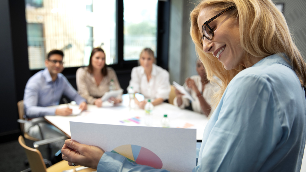Eine lächelnde Frau mit blonden Haaren und Brille steht im Vordergrund eines modernen Konferenzraums und hält ein Blatt mit einem bunten Tortendiagramm in der Hand. Im Hintergrund sitzen vier unscharf dargestellte Personen (drei Frauen, ein Mann) an einem Tisch und hören aufmerksam zu. Auf dem Tisch liegen Wasserflaschen und bunte Diagramme.
Die Szene vermittelt eine positive und professionelle Atmosphäre in einem Geschäftstreffen oder Workshop.